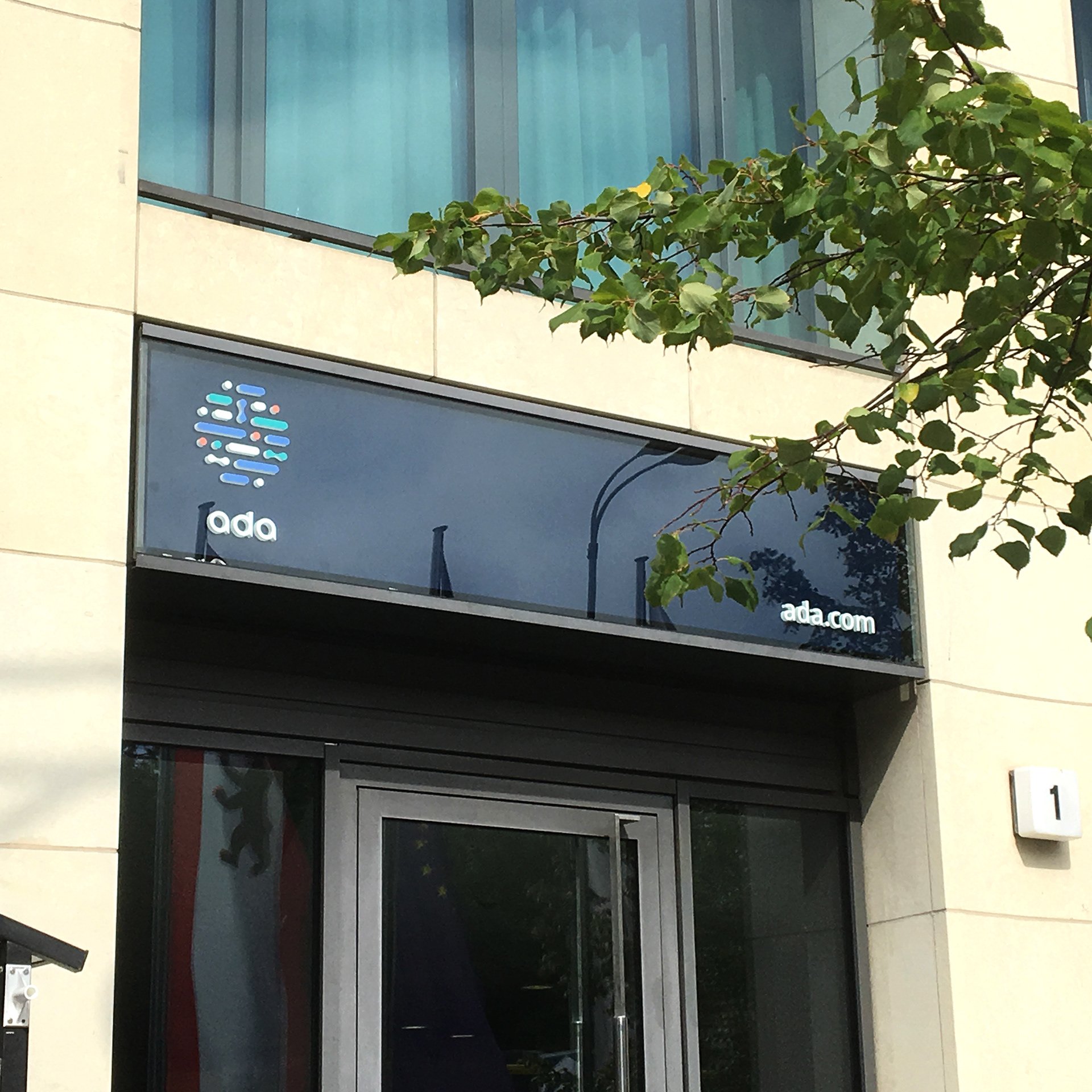 Berlin HQ entrance — illuminated ‘ada’ wordmark and brand mark mounted above the door, ‘ada.com’ decal on the glass
