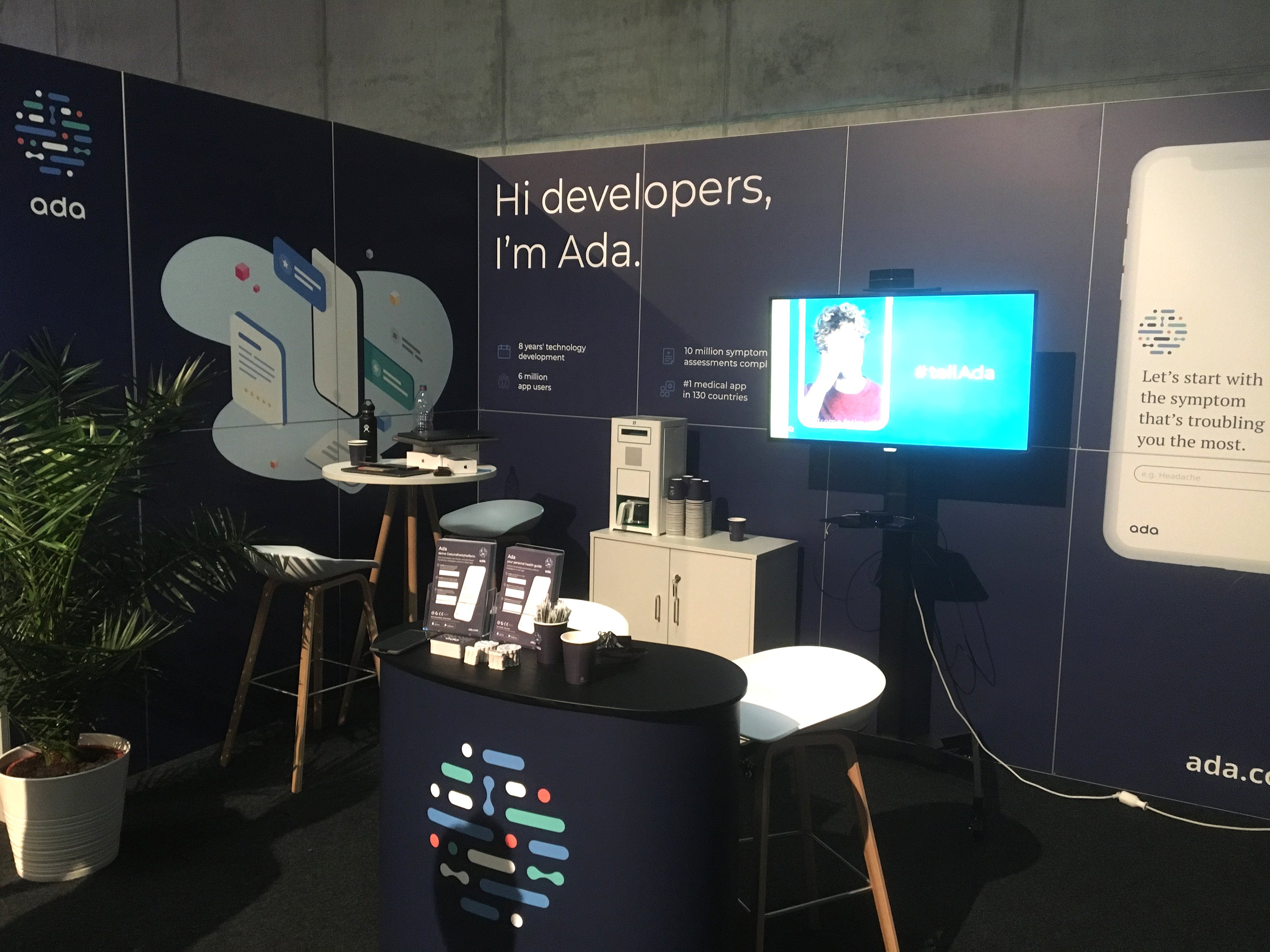 Conference booth at a developer event — ‘Hi developers, I’m Ada’ back wall with the illustrated brand pattern, headline stats, screens, bar stools and a branded counter