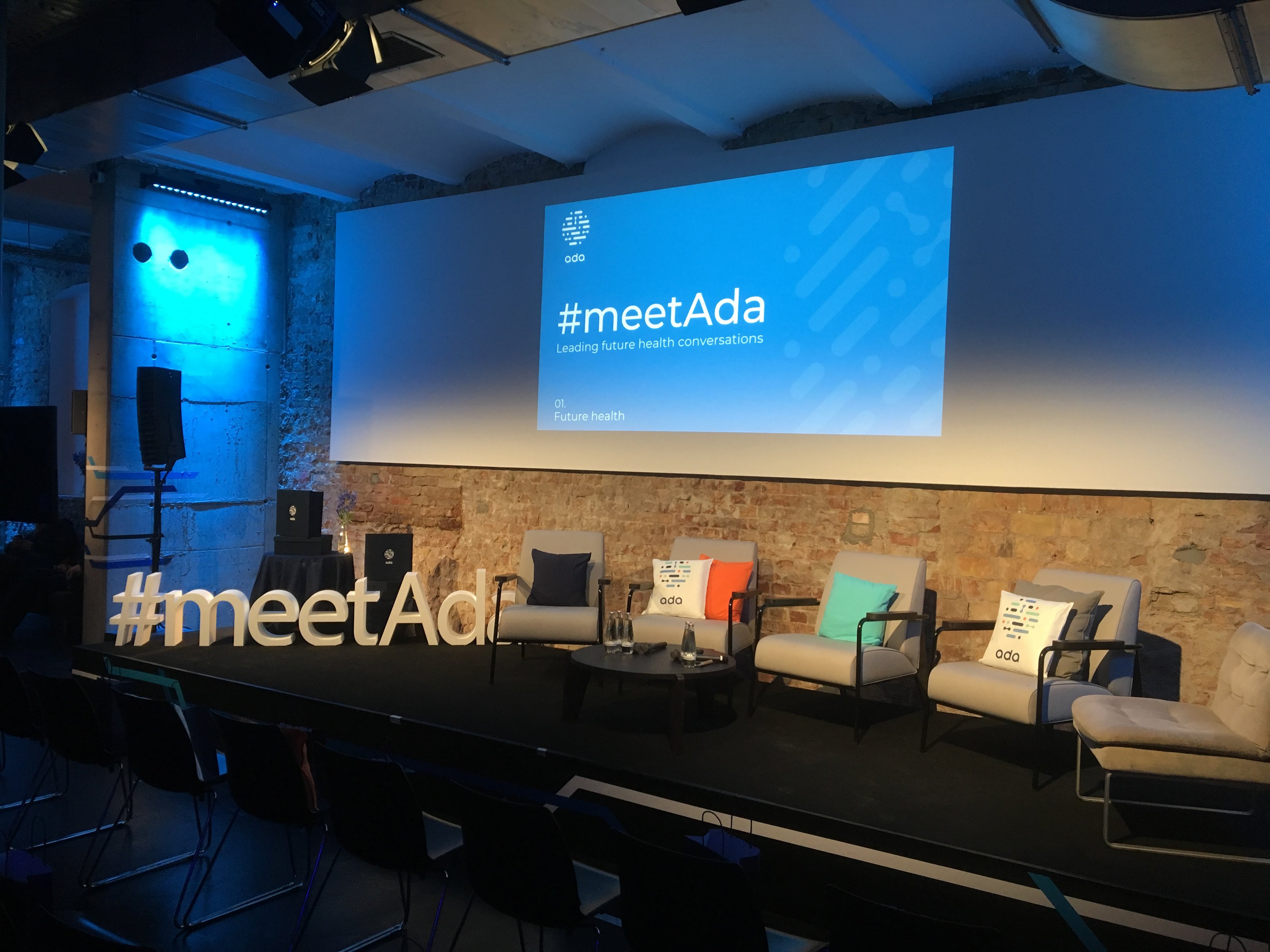#meetAda event stage — freestanding ‘#meetAda’ letterforms on a brick-walled stage with branded cushions, ‘#meetAda — Leading future health conversations’ projected behind