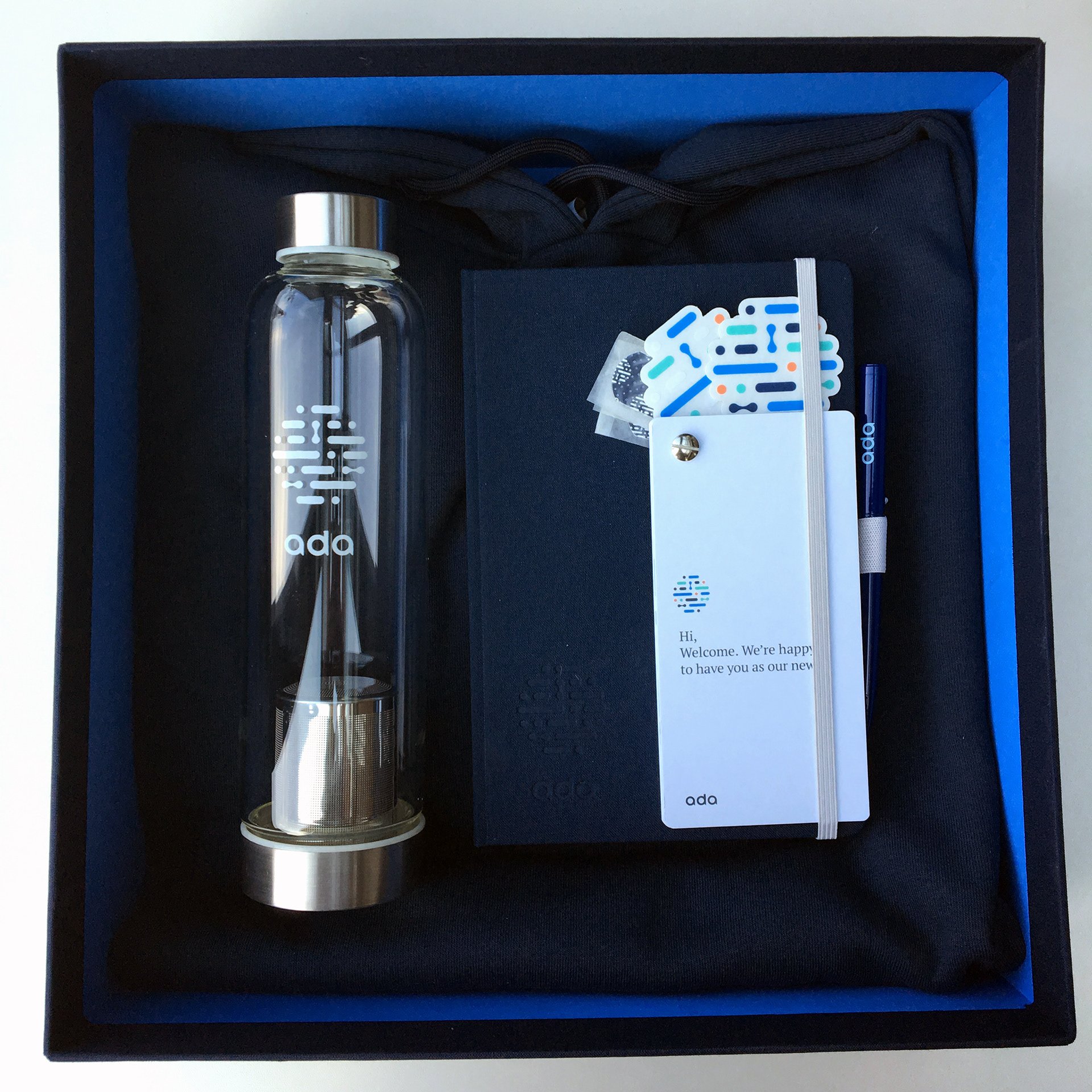 Onboarding box opened — water bottle, branded notebook, hoodie, ‘Hi, Welcome’ card, pen and sticker laid out inside the navy box