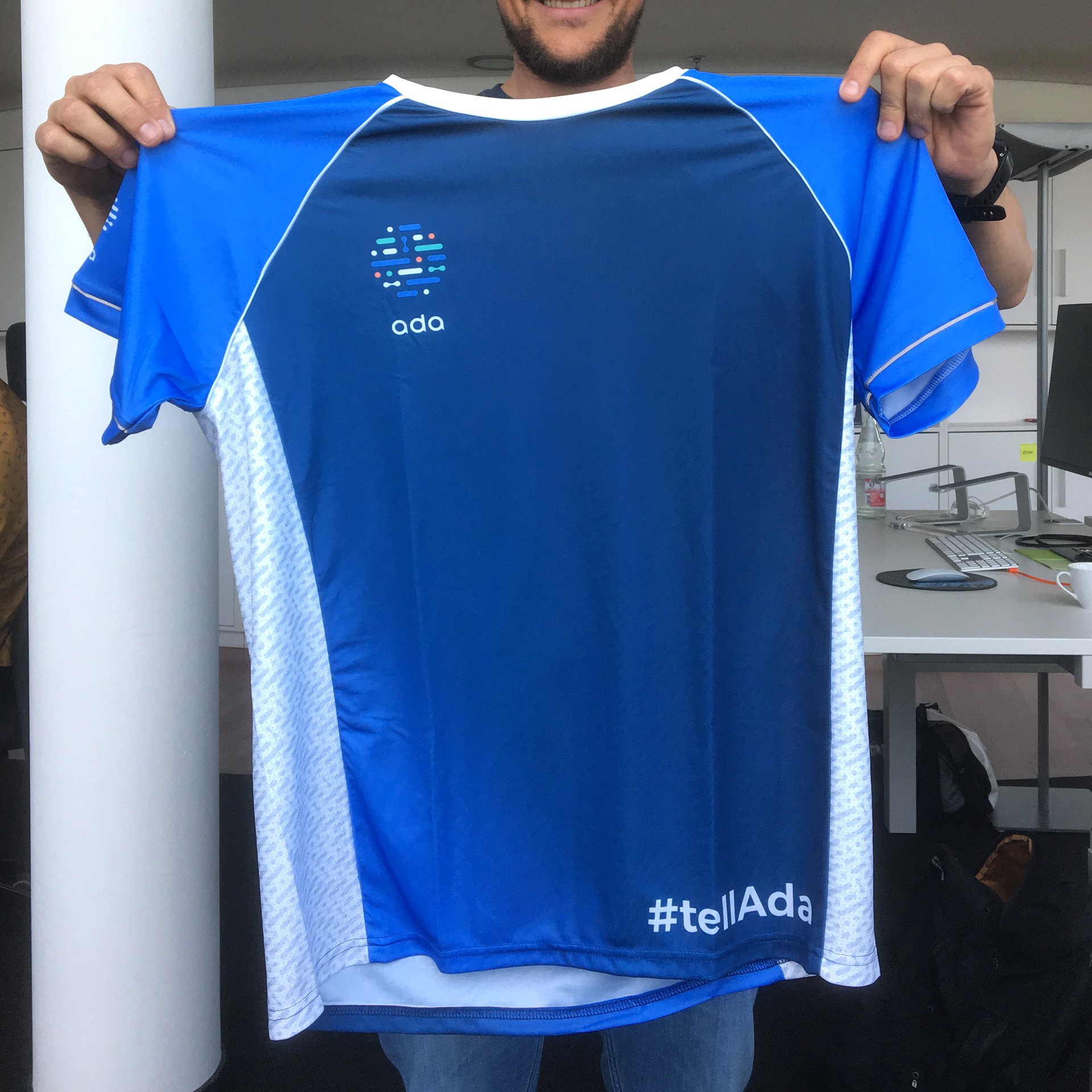 Ada running shirt held up in the office — blue jersey with the Ada logo on the chest and ‘#tellAda’ on the lower back