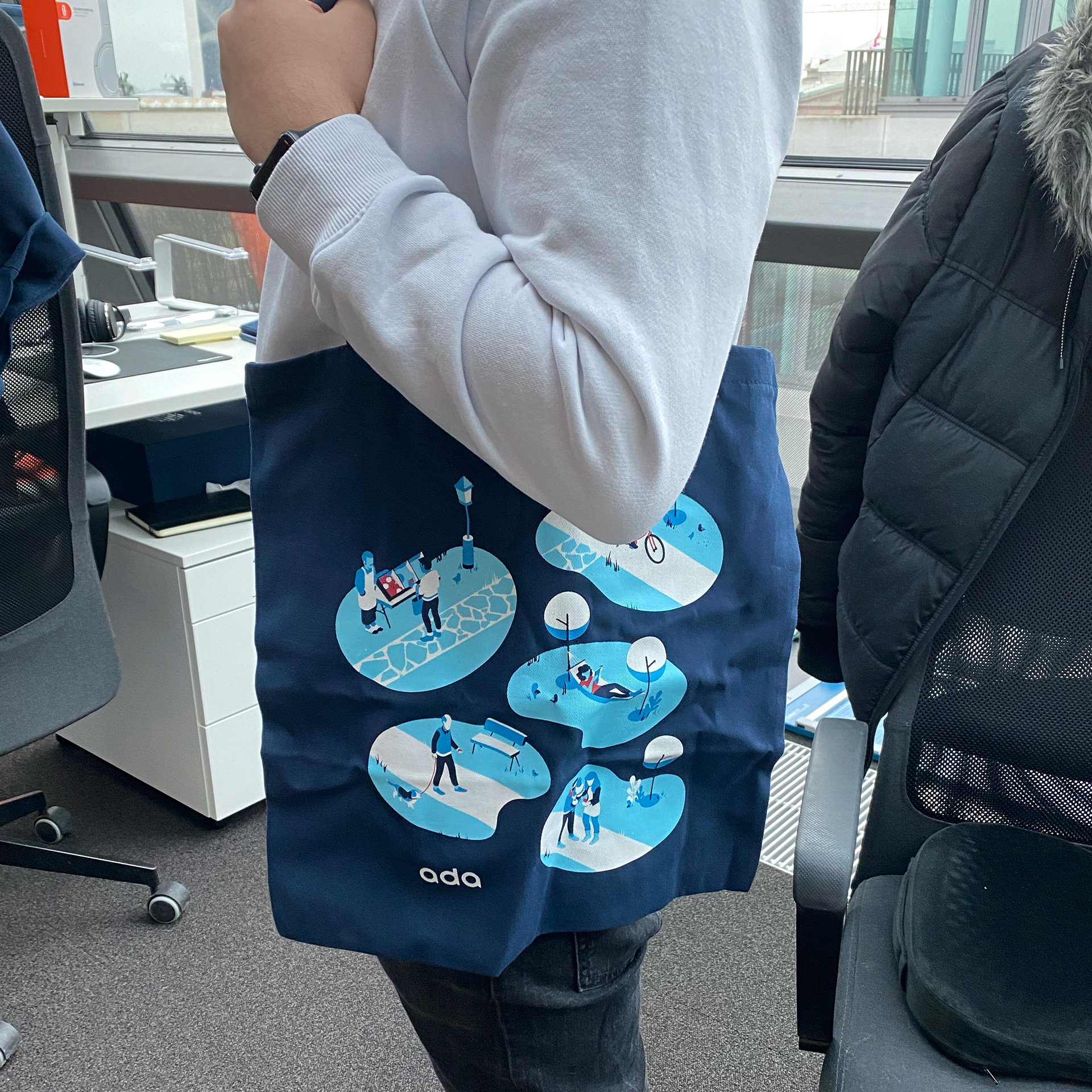 Ada-branded tote bag carried in the office — navy canvas with an illustrated brand-system pattern of small scenes and the Ada wordmark