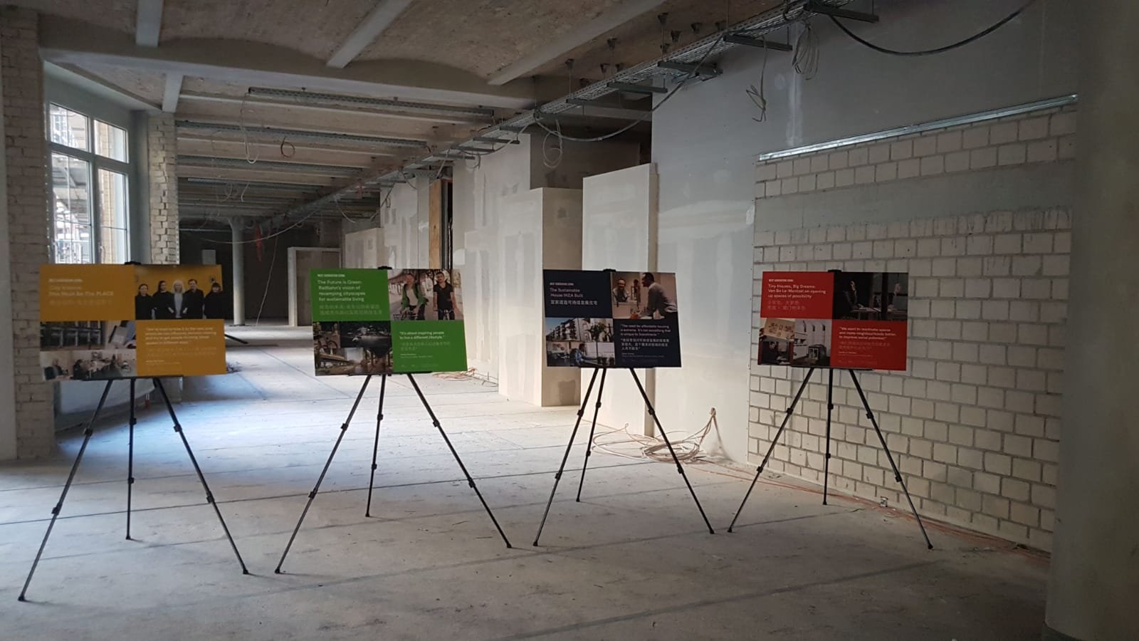 Next Generation Living launch event — four colour-coded A1 information panels on tripod stands in an industrial Berlin venue, showing the yellow, green, navy and red-orange topic-colour system installed in the space